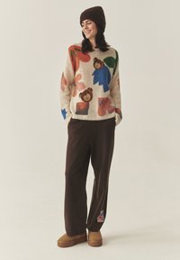 Pleteni pulover z barvitimi cvetličnimi in medvedjimi vzorci, bež podlaga; rjave hlače z visoko pasom; rjavi gležnjarji z teksturirano površino.