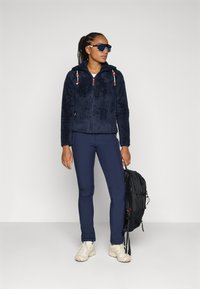 Marinblå fleecejacka med dragkedja, tan accenter och huva; kombinerad med marinblå leggings och ljusa skor; håller en svart ryggsäck.