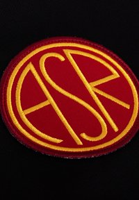 Patch circolare rosso ricamato con le lettere "ASR" in giallo, su uno sfondo di tessuto nero. Bordato con cuciture rosse.