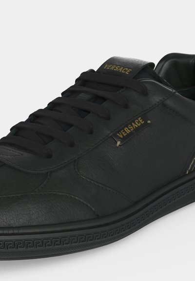 Μαύρο δερμάτινο αθλητικό παπούτσι Versace με μαύρα κορδόνια, χρυσό λογότυπο Versace στη πλευρά και στη γλώσσα, και διακοσμημένη μαύρη ελαστική σόλα.