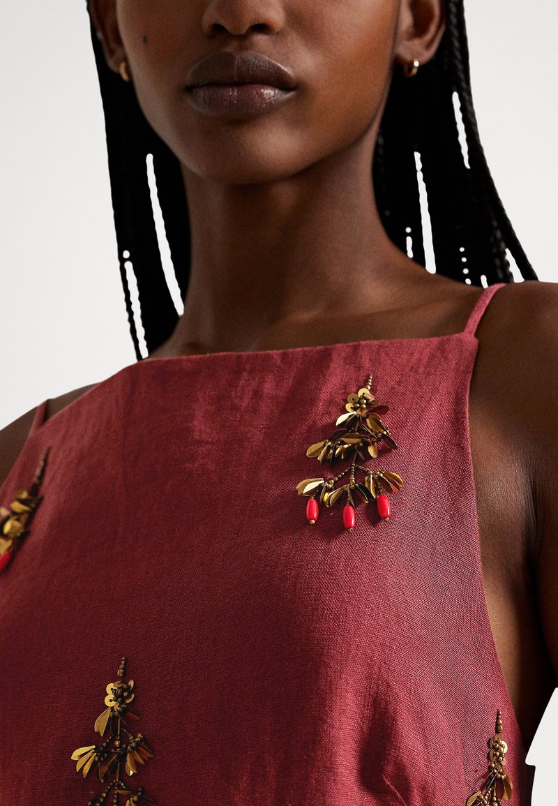 Nahaufnahme einer Person, die ein ärmelloses dunkelrotes Kleid trägt, das mit goldenen und roten Blatt- und Beerenverzierungen geschmückt ist.