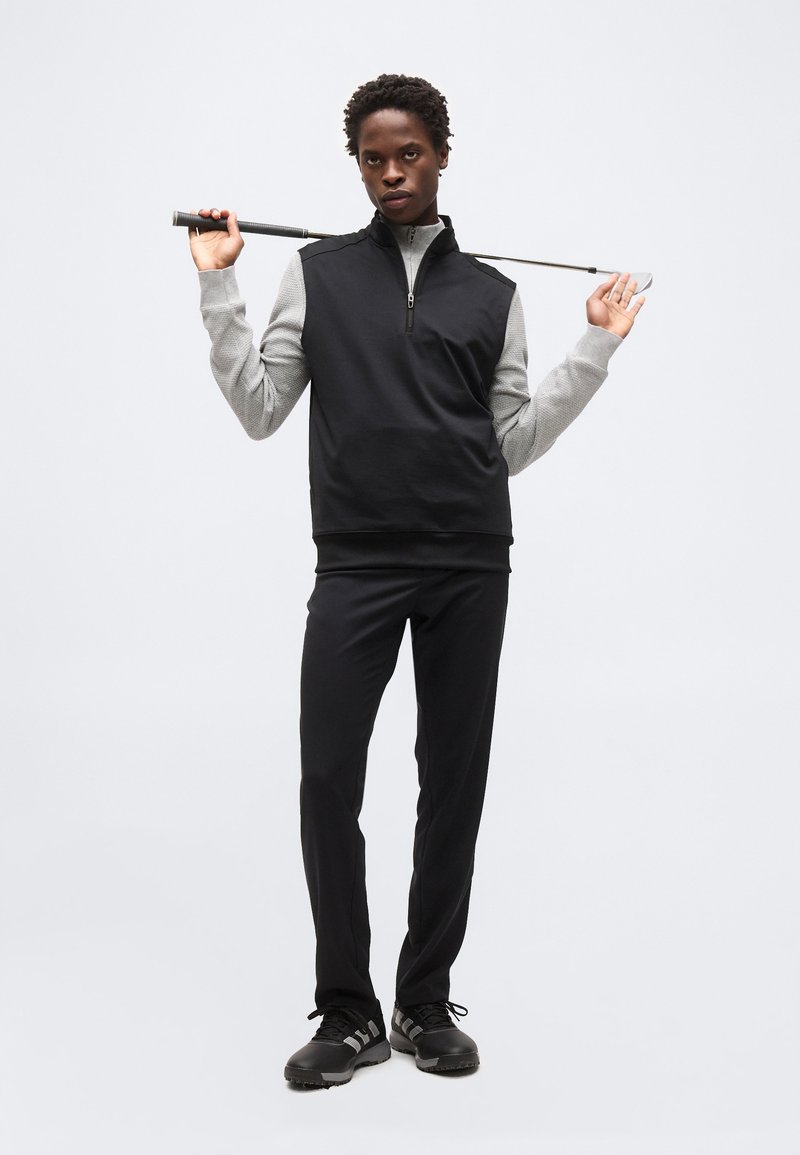 Personne tenant un club de golf derrière le cou, portant un gilet sans manches noir sur une chemise grise à manches longues, un pantalon noir et des chaussures noires, debout devant un fond blanc.