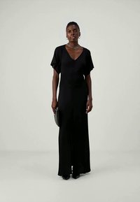 Μαύρο μακρύ φόρεμα με V-λαιμόκοψη, κοντές φουσκωτές μανσέτες και ρέουσα σιλουέτα. Συνδυασμένο με μαύρη τσάντα και γόβες με μυτερή μύτη.