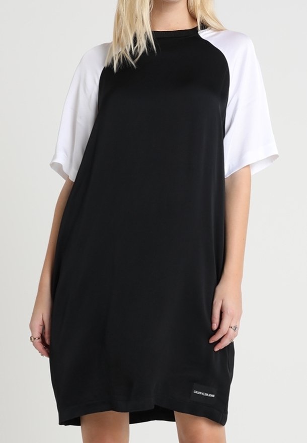 Femme portant une robe noire ample avec des manches raglan blanches courtes, debout devant un fond clair uni.