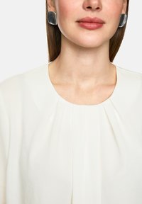 Silberne quadratische Ohrringe mit glatter Oberfläche; Modell trägt eine cremefarbene Bluse mit auffälligen Falten am Ausschnitt, wobei der Schlüsselbeinbereich sichtbar ist.