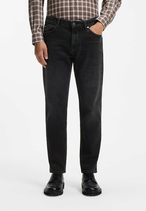Mann, der schwarze Jeans, schwarze Schuhe und ein braunes kariertes Langarmhemd trägt, mit einer Hand in der Tasche.