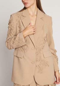Blazer bej cu aplicații florale texturate pe mâneci și față, având închidere cu un singur buton și rever în formă de palmă. Material neted, structurat.