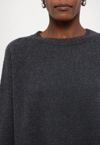 Gros plan d'une personne portant un pull en maille gris foncé à encolure ronde et des boucles d'oreilles argentées en forme d'anneau.