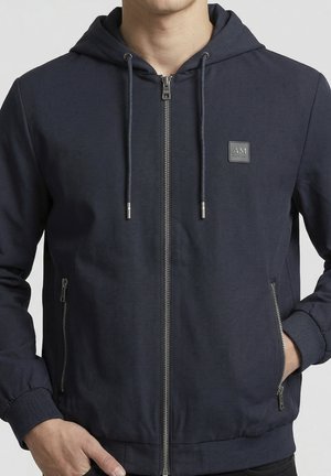 Man draagt marineblauwe capuchontrui met metalen ritsen en AM-logo op de borst, handen in de zakken.