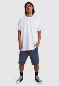 Weißes Baumwoll-T-Shirt, locker geschnitten, kombiniert mit dunkelgrauen Shorts mit Taschen. Schwarze High-Top-Sneaker, einfarbig, mit weißen Schnürsenkeln.
