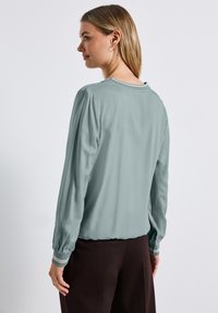 Blouse bleu clair avec de longues manches froncées et un col rond. Présente des rayures contrastantes sur les poignets, une texture lisse et une coupe décontractée.