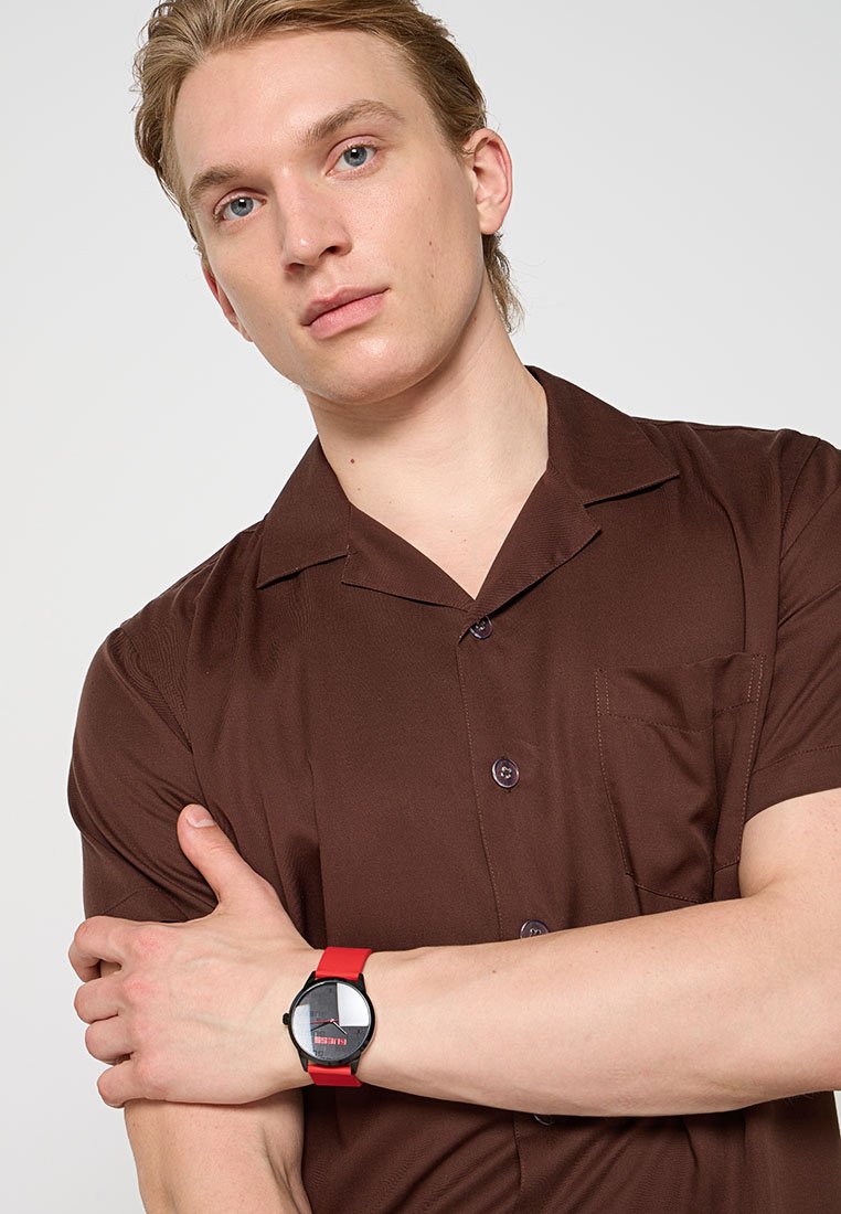 Joven con una camisa de manga corta marrón y los brazos cruzados, que lleva un reloj de pulsera negro y rojo en la muñeca izquierda.