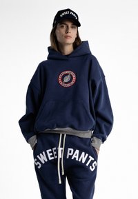 Sweat à capuche marine avec logo brodé en design circulaire, associé à un pantalon de survêtement assorti portant le texte "SWEET PANTS" en blanc gras. Accents gris.