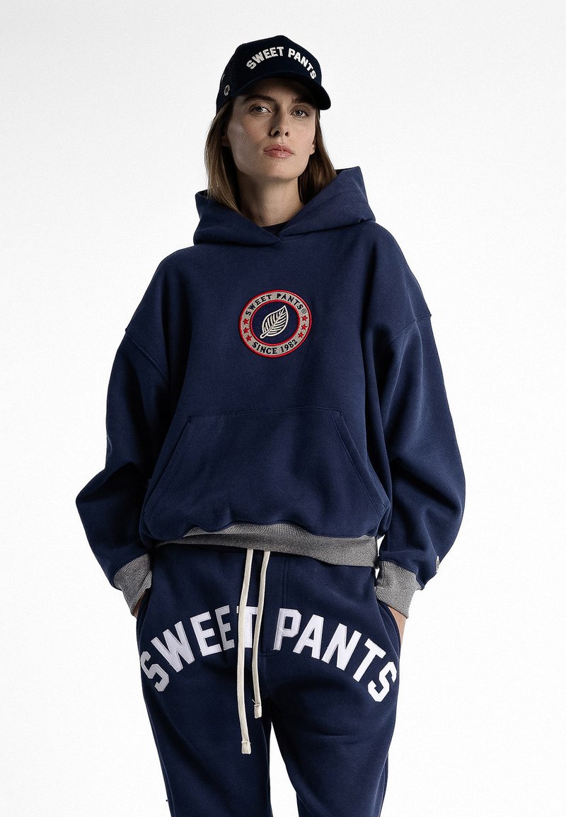 Sweat à capuche marine avec logo brodé en design circulaire, associé à un pantalon de survêtement assorti portant le texte "SWEET PANTS" en blanc gras. Accents gris.