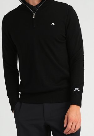 Mand iført en sort langærmet quarter-zip sweater med logo på bryst og ærme, kombineret med mørke bukser, står med hånden i lommen.