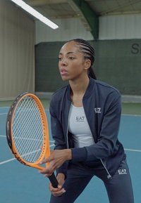 Tenisový hráč v tmavej zipsovej bundě a bielom tričku, s oranžovou raketou, pripravujúci sa na podanie na modrom kurte.