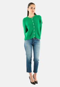 Cardigan verde lavorato a maglia con scollo rotondo, chiusura a bottoni e vestibilità ampia, abbinato a jeans skinny blu chiaro e tacchi neri.