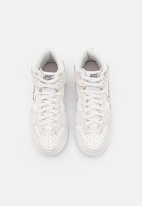 Vaaleanbeigeja ja valkoisia nahkaisia korkeita tenneriä, joissa on rei'itetty kärki, litteät nauhat ja vaaleanpunainen Nike-logo sivussa.