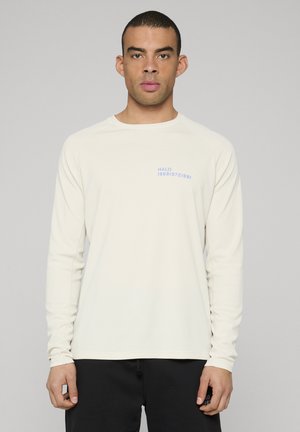 Beiges Langarm-Sportshirt mit strukturiertem Muster und blauem Aufdruck "HALO 196819721981" auf der linken Brustseite.