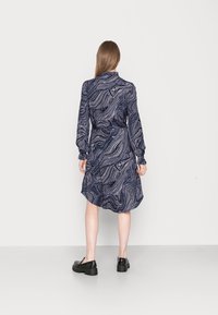 Pitkähihaiset mekko tummansinistä väriä valkoisella kiemurtelevalla kuviolla, vyötetty vyötäröltä ja kaareva helma. Yhdistetty mustiin kenkiin.