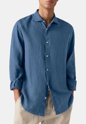 Uomo che indossa una camicia blu a maniche lunghe con bottoni e polsini arrotolati e pantaloni beige, mani in tasca, su uno sfondo semplice.