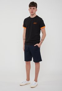 T-shirt nera a maniche corte con dettagli arancioni sulle maniche, abbinata a pantaloni corti blu navy e sneakers bianche, in piedi contro uno sfondo semplice.