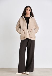 Giacca in pile beige con grandi tasche, chiusura a bottoni e accenti grigi a contrasto sulle spalle. Abbinata a pantaloni scuri e scarpe chiare.