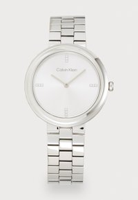 Montre en acier inoxydable argenté avec un boîtier rond, un cadran blanc, des marqueurs d'heure minimalistes et un bracelet en maillons métalliques. Logo de la marque placé sur le cadran.