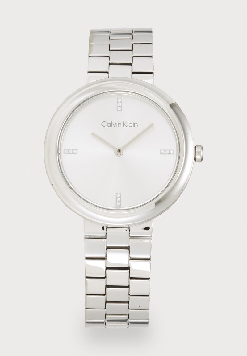 Montre en acier inoxydable argenté avec un boîtier rond, un cadran blanc, des marqueurs d'heure minimalistes et un bracelet en maillons métalliques. Logo de la marque placé sur le cadran.