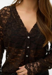 Črna čipkasta bluza z V-izrezom, dolgimi rokavi in majhnimi dekorativnimi gumbi. Tkanina ima cvetlični vzorec s prosojno teksturo.