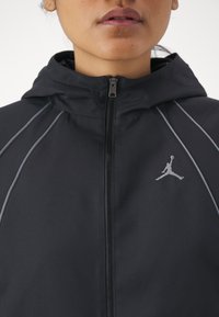 Svart huva jacka med silverfärgade sömmar och dragkedja. Har en Jumpman-logotyp på vänster bröst.