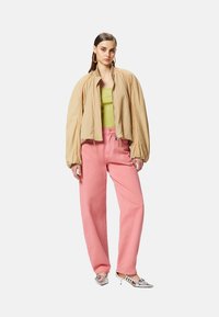Giacca bomber beige, top verde aderente e jeans rosa larghi. Scarpe con tacco argento valorizzano l'outfit, presentando dettagli e texture uniche.