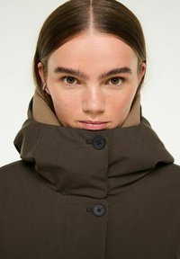Bruine buitenjas met een hoge kraag, voorzien van twee zwarte knopen en een gladde textuur. De binnenkraag is beige.