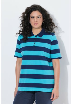 Kvinde med langt krøllet hår iført en blå og turkisstribet poloshirt og mørke bukser, stående mod en hvid baggrund.