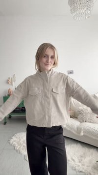 Junge Frau trägt eine beige Jacke und schwarze Hose, steht in einem hellen Wohnzimmer mit einem weißen Sofa und einem Kronleuchter darüber.