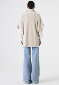 Person with dark hair in a bun wearing a beige oversized sweater over a white blouse and wide-leg light blue jeans, standing facing away.