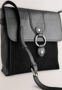 Sac à bandoulière en cuir et daim noir avec fermeture à rabat, détail anneau argenté et sangle ajustable sur un fond clair.