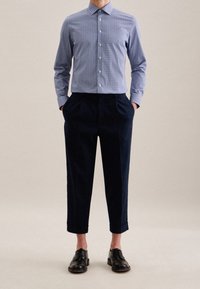 Muž v modré vzorované košili na knoflíky, tmavomodrých zkrácených kalhotách s manžetami a černých kožených obuvích, stojící s rukama v kapsách.