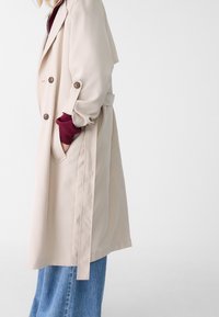 Trench-coat beige à double boutonnage avec de grands boutons, une ceinture à la taille et des poches. Il présente une texture lisse et une coupe décontractée, porté par-dessus un haut bordeaux.