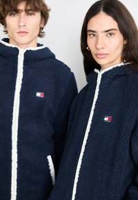 Mornar plavi flis jakne na zip s bijelim detaljima, s logotipom Tommy Jeans. Tekstura je pahuljasta i mekana, s dizajnom kapuljače.