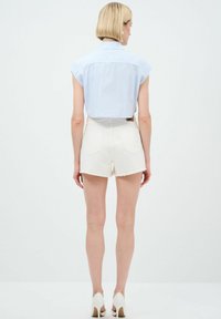 Camicetta azzurra chiara corta con maniche corte, abbinata a pantaloni shorts in denim bianchi con bordo sfrangiato e tasche posteriori. Sfondo trasparente, colori neutri.