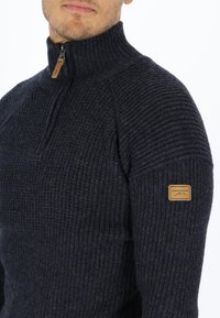 Navy ribbed knit sweater with a high collar and a quarter zip. Features a brown logo patch on the left shoulder. Soft texture.