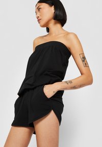 Sort strapless jumpsuit lavet af strækbart materiale, med en elastisk halsudskæring, lommer og en løstsiddende pasform med sidesplit.