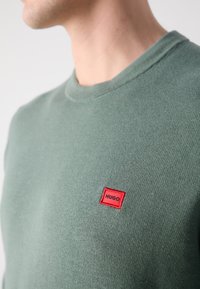 Green knit sweatshirt with a crew neckline and a small red rectangular logo patch labeled "HUGO" on the left side.