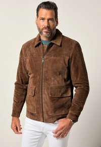 Un hombre con barba posa con confianza, vistiendo una chaqueta de ante marrón sobre una camisa verde azulado, complementada con jeans blancos. Fondo simple.