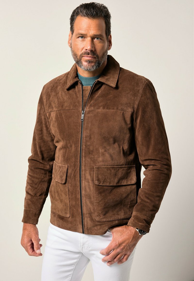 Un hombre con barba posa con confianza, vistiendo una chaqueta de ante marrón sobre una camisa verde azulado, complementada con jeans blancos. Fondo simple.