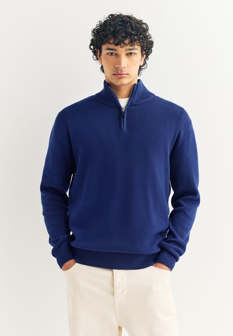 Joven con cabello rizado, vistiendo un suéter azul marino con cierre parcial y pantalones beige claros, de pie con las manos en los bolsillos.