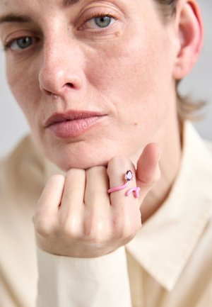 Anillo rosa con un diseño ondulado y un acento de gema púrpura, llevado en un dedo. Textura de la piel y uña natural visibles en primer plano.