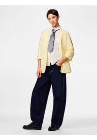 Jeune personne debout, main dans la poche, portant un blazer jaune, une chemise blanche, une cravate rayée, un large jean bleu foncé et des chaussures noires.