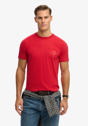 Rotes Baumwoll-T-Shirt mit kurzen Ärmeln und einer Brusttasche mit aufgedrucktem Text. Kombiniert mit blauen Jeans und einem karierte Hemd, das in der Taille gebunden ist.